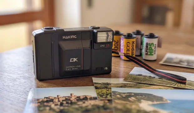 La compatta Haking CFE 35 con il cinturino nero e rosso, su un tavolo di legno, tra rullini 35 mm e stampe di paesaggi e borghi