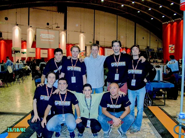 Nove ragazzi in posa in un padiglione dello SMAU, molti con la maglietta del gruppo SOOL e il pass al collo; alle spalle le postazioni della gara e gli stand della fiera